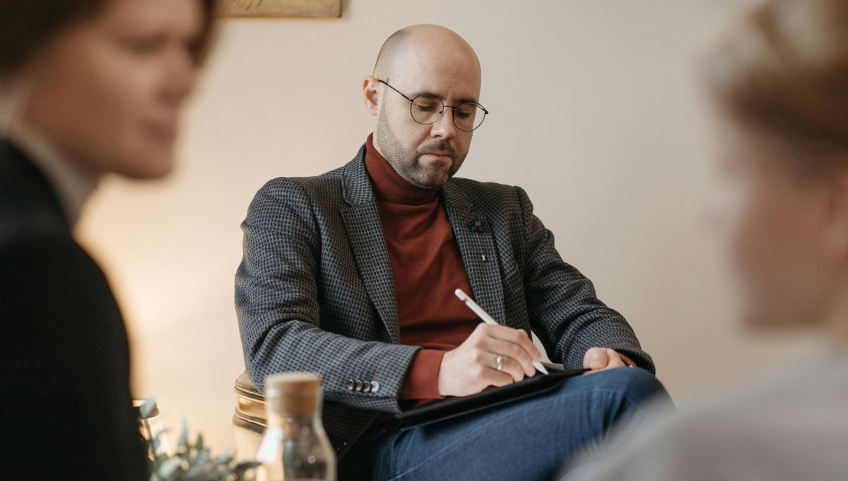 Ein Psychotherapeut auf einem Stuhl mit einem Stift in der Hand, Vor ohm unterhalten sich zwei Menschen.